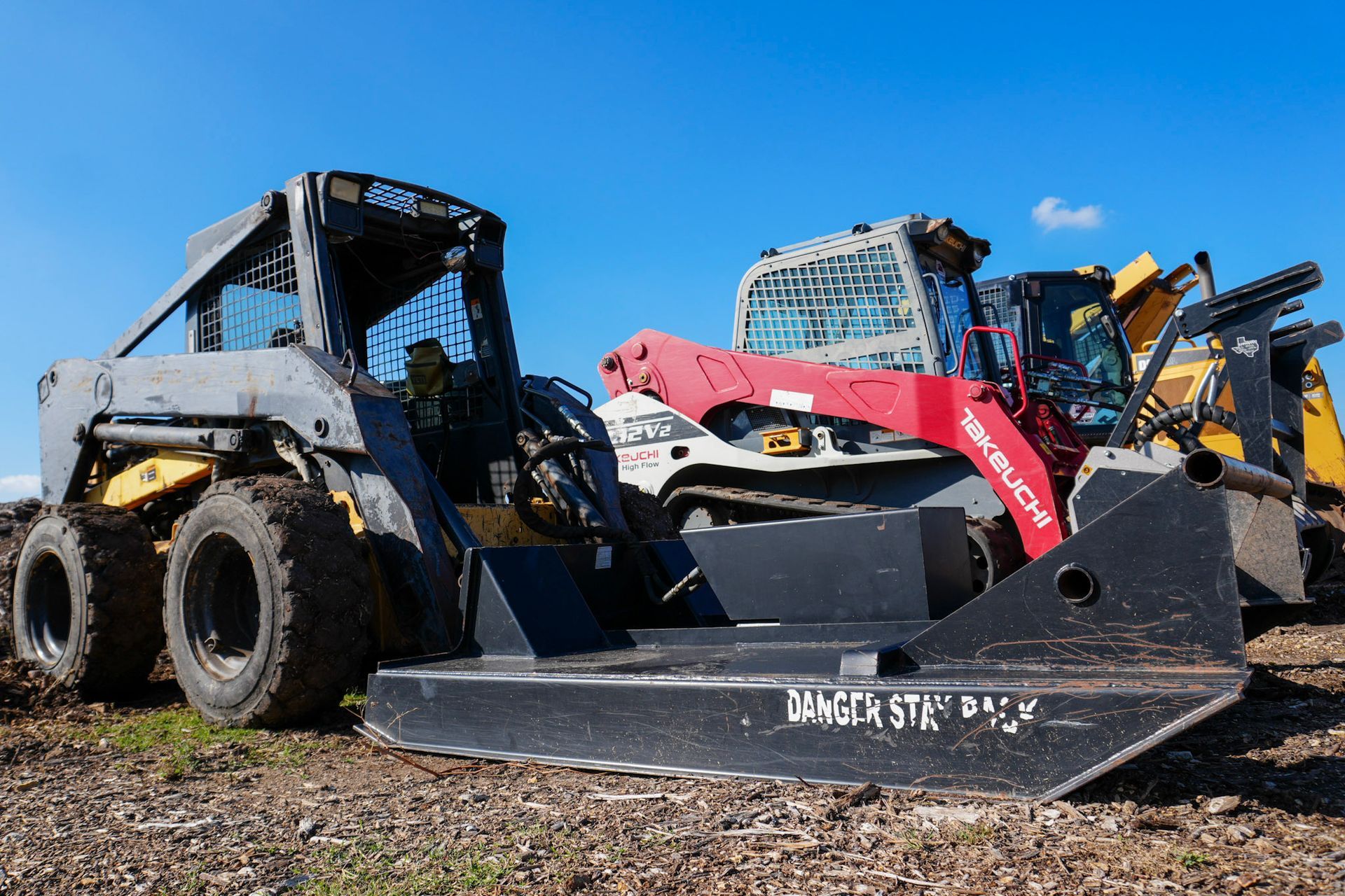 Two skid steer loaders, one gray and one red, parked on a dirt lot under a clear blue sky.