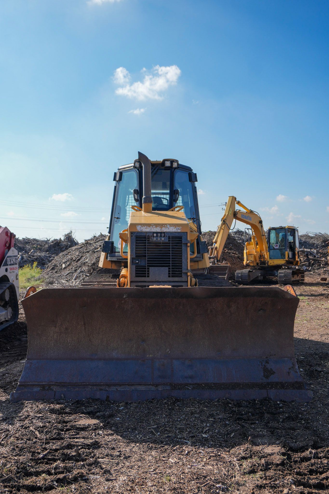 A yellow bulldozer sits in the foreground on a dirt lot, with a yellow excavator and a red machine parked nearby.