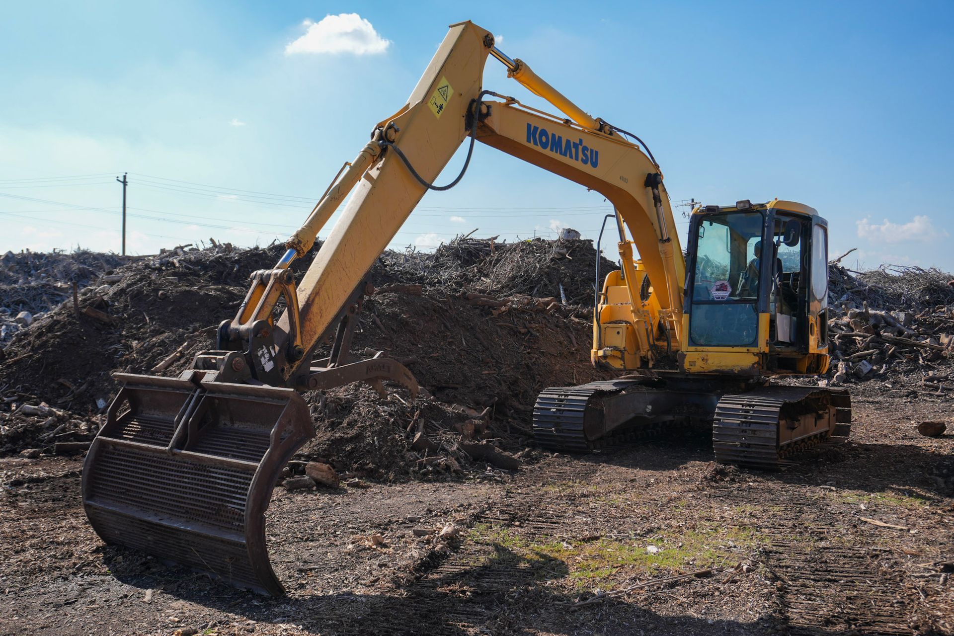 A yellow Komatsu excavator with a rake-style bucket operates at a large debris site under a bright blue sky.