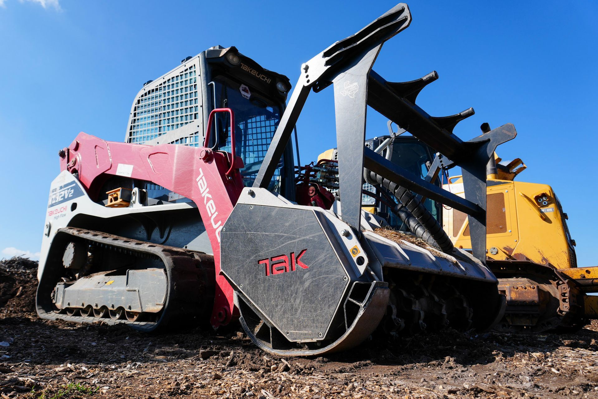 A red and white skid-steer loader with a mulching attachment sits on dirt ground under a clear blue sky.