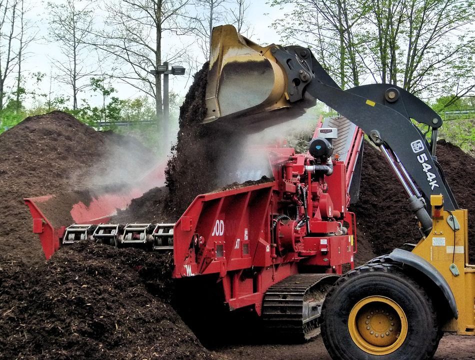 A red wood chipper being loaded by a yellow and black loader, in a yard with mulch piles.