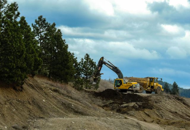 An excavator and dump truck working on a hillside. Trees and cloudy sky in the background.