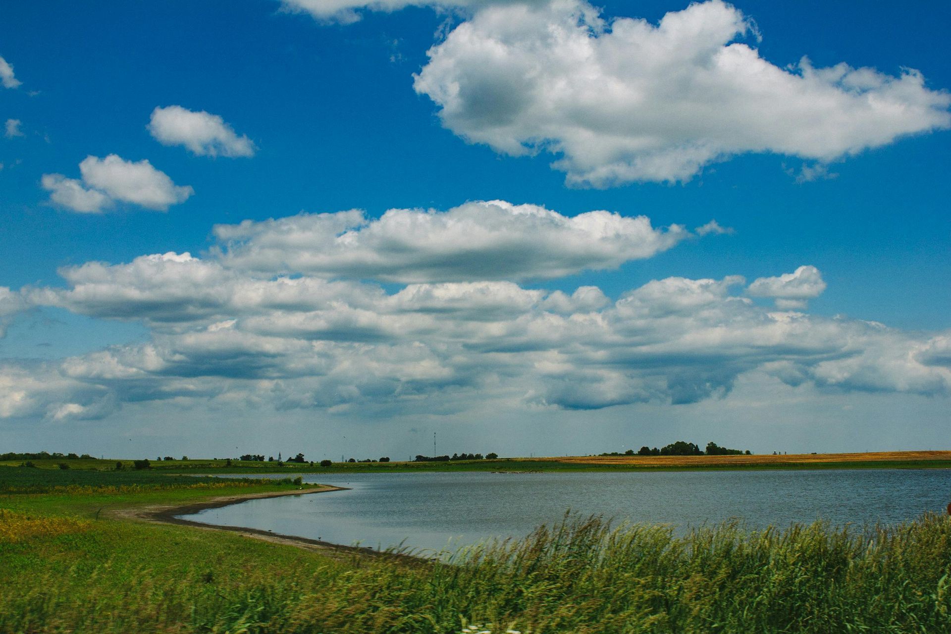 Blue sky with white clouds over a lake with green fields and dry grass on the shore.