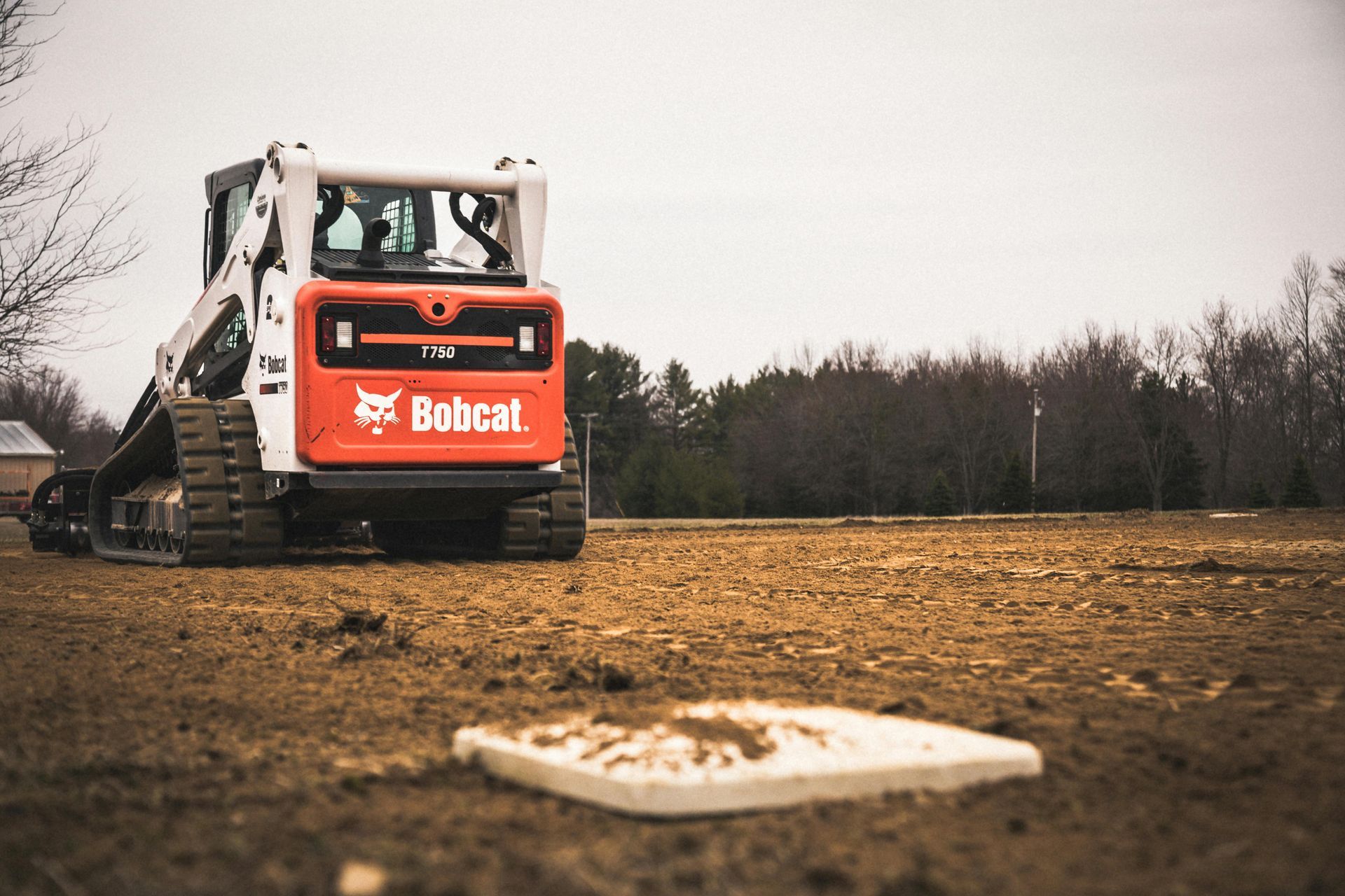 Bobcat track loader on a dirt baseball field near home plate, cloudy day.