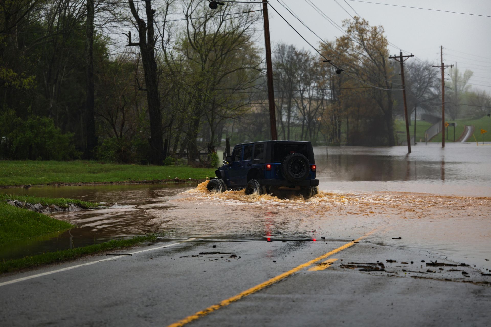 A dark-colored Jeep drives through a flooded road, creating a splash as it moves across the water.