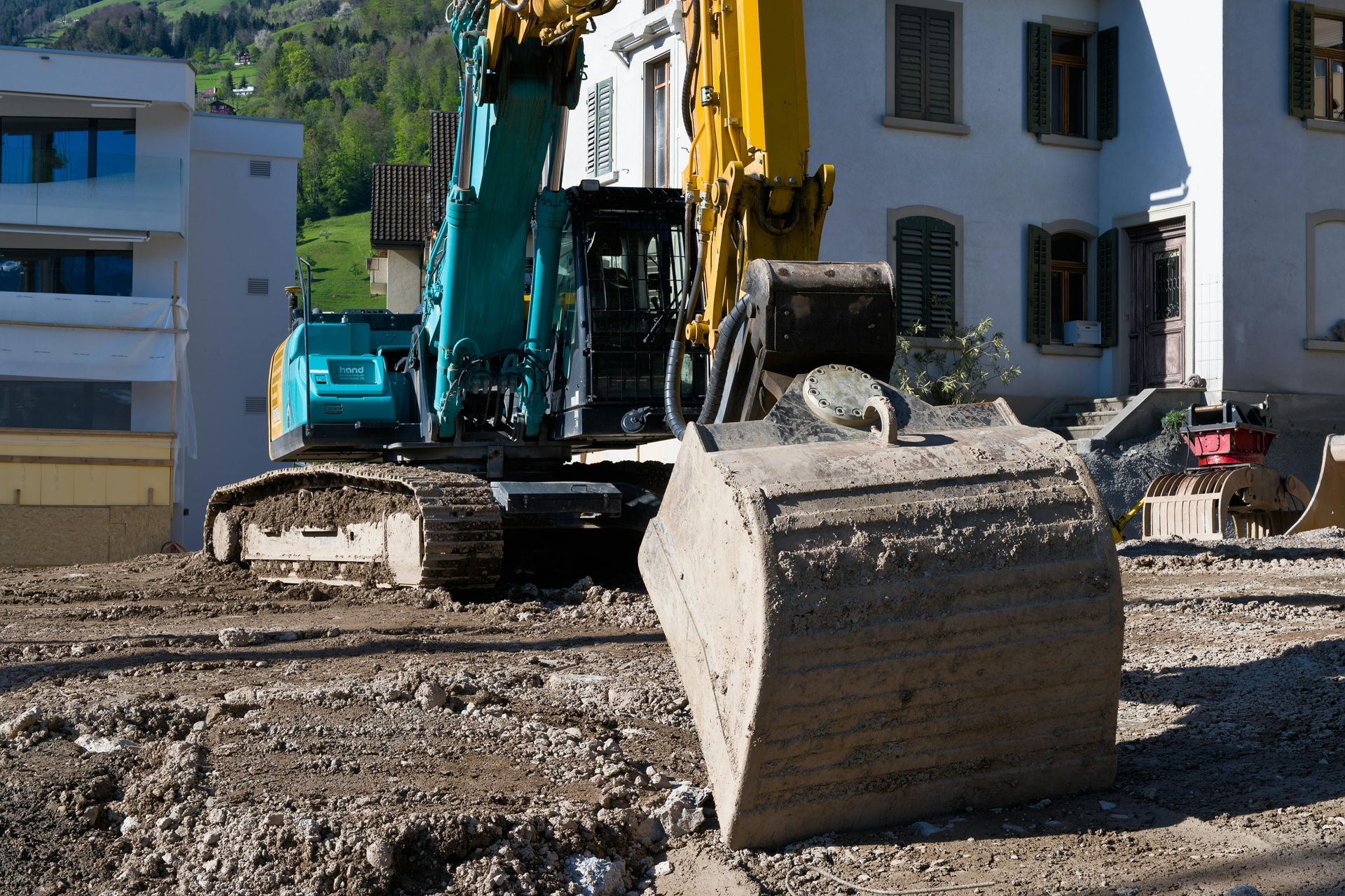A construction site with an excavator in front of a building. The excavator is teal and yellow.