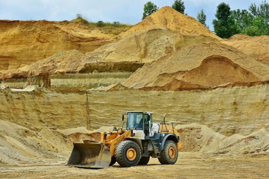 Yellow front-end loader in a sand pit. Tan and brown mounds of dirt and trees in the background.