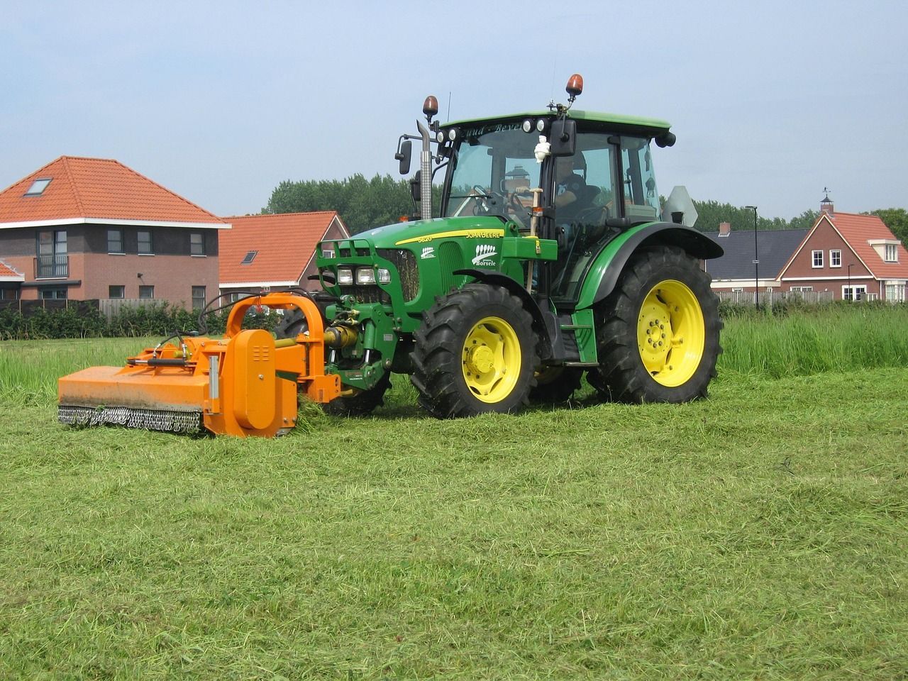 Green tractor with orange mower cutting grass in a field, houses in the background.