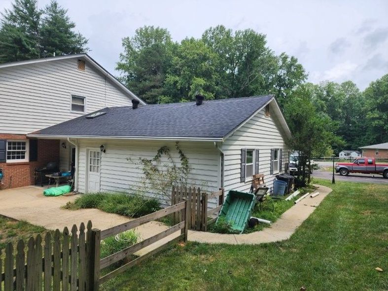 A side view of a white, single-story house with a new dark gray shingle roof, a concrete patio, and a wooden fence.