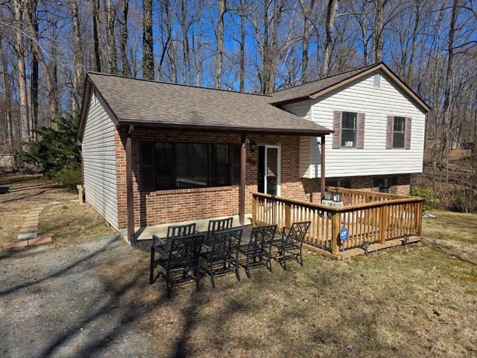 A two-story house with a brick front, tan siding, and a wooden deck featuring a row of chairs in a wooded setting.
