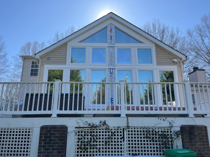 A sunny house exterior featuring a large wall of windows, a white deck railing, and a lattice-covered brick foundation.