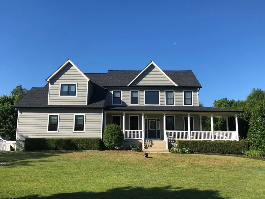 Two-story light gray house with a dark roof and large front porch, set against a clear blue sky and green lawn.