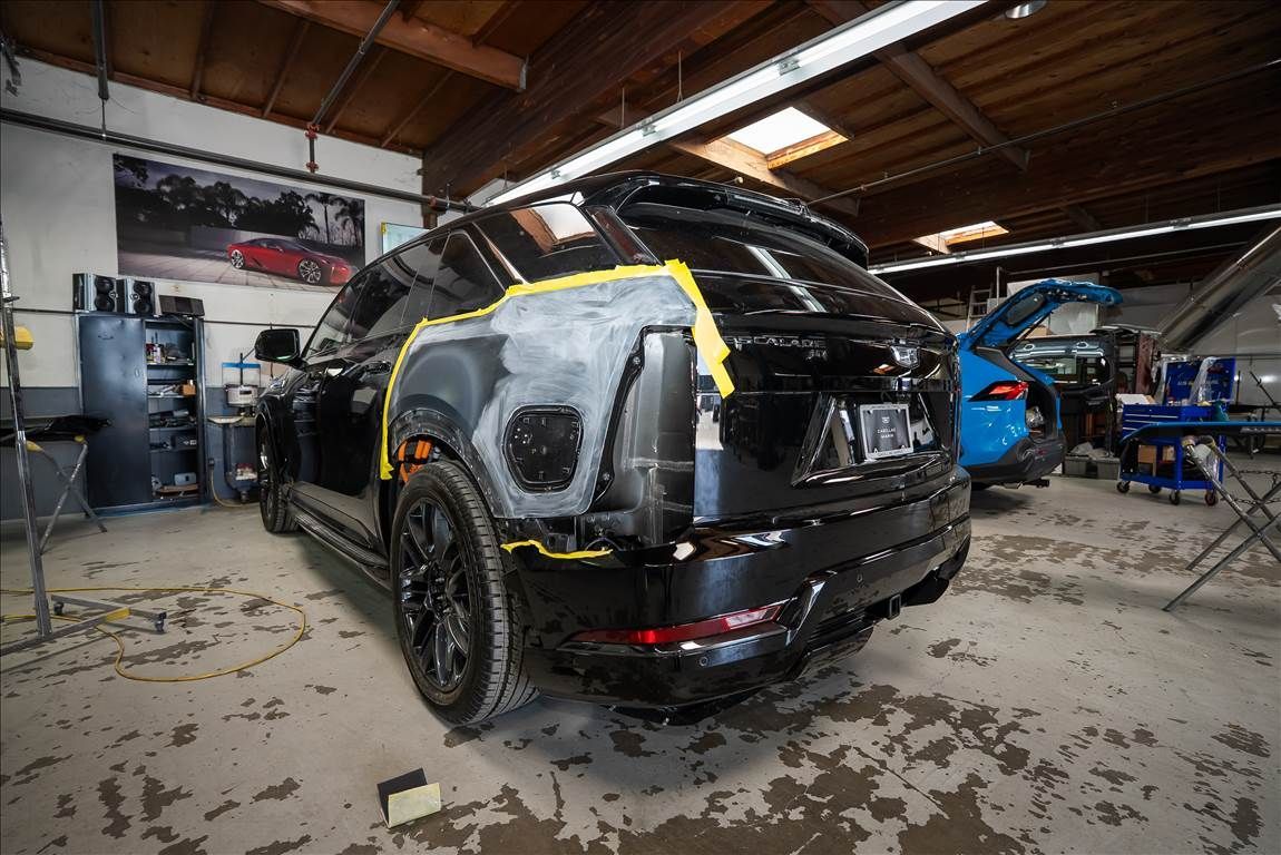 Black SUV being repaired in a garage; bodywork completed, taped edges, and a blue vehicle is visible in the background.