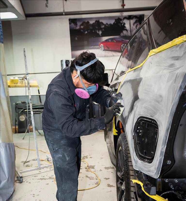 Auto body worker sanding a car's panel while wearing a respirator and gloves in a garage.