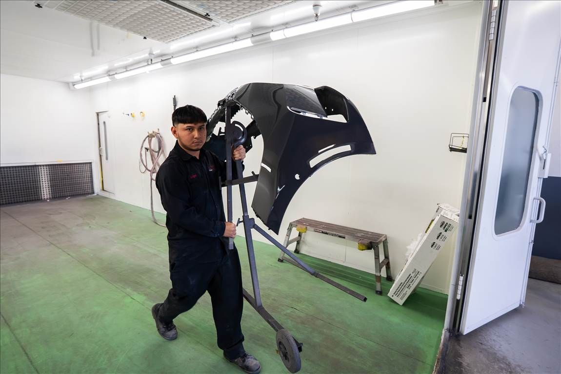 Man in a garage carries a black car bumper on a stand. White walls, green floor, and fluorescent lights.