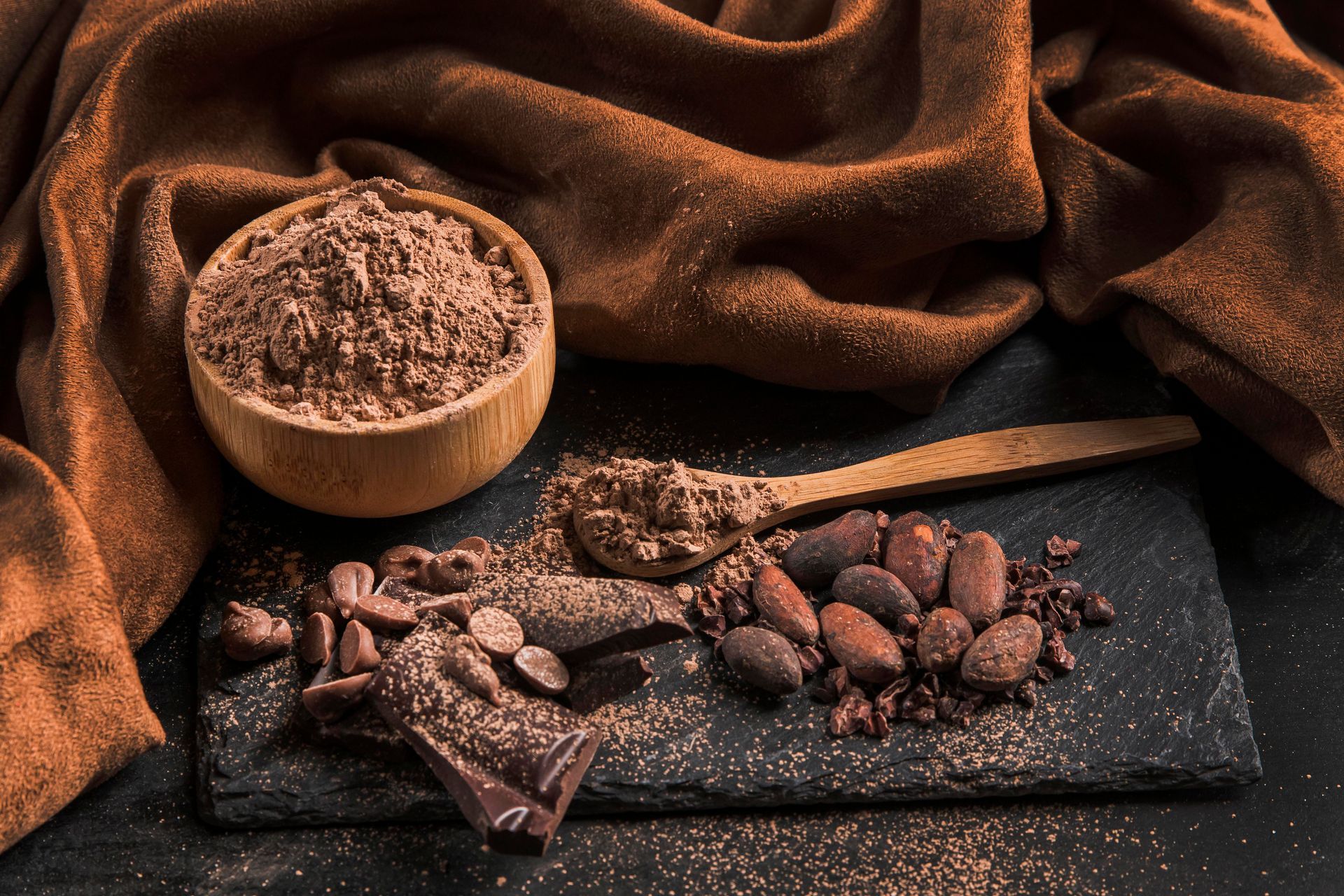 A bowl of cocoa powder next to a pile of cocoa beans and a wooden spoon.