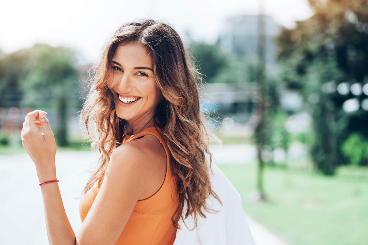Eine Frau in einem orangefarbenen Tanktop lächelt und hält eine weiße Tasche.