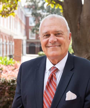 Older white man in suit and tie smiles in front of a brick building.