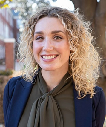 Woman with curly blonde hair, smiling, wearing a dark blazer and olive green blouse, outdoors.