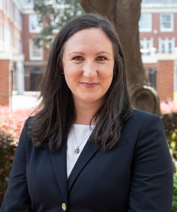Woman in dark blazer and white shirt smiles, outdoors, in front of a building.