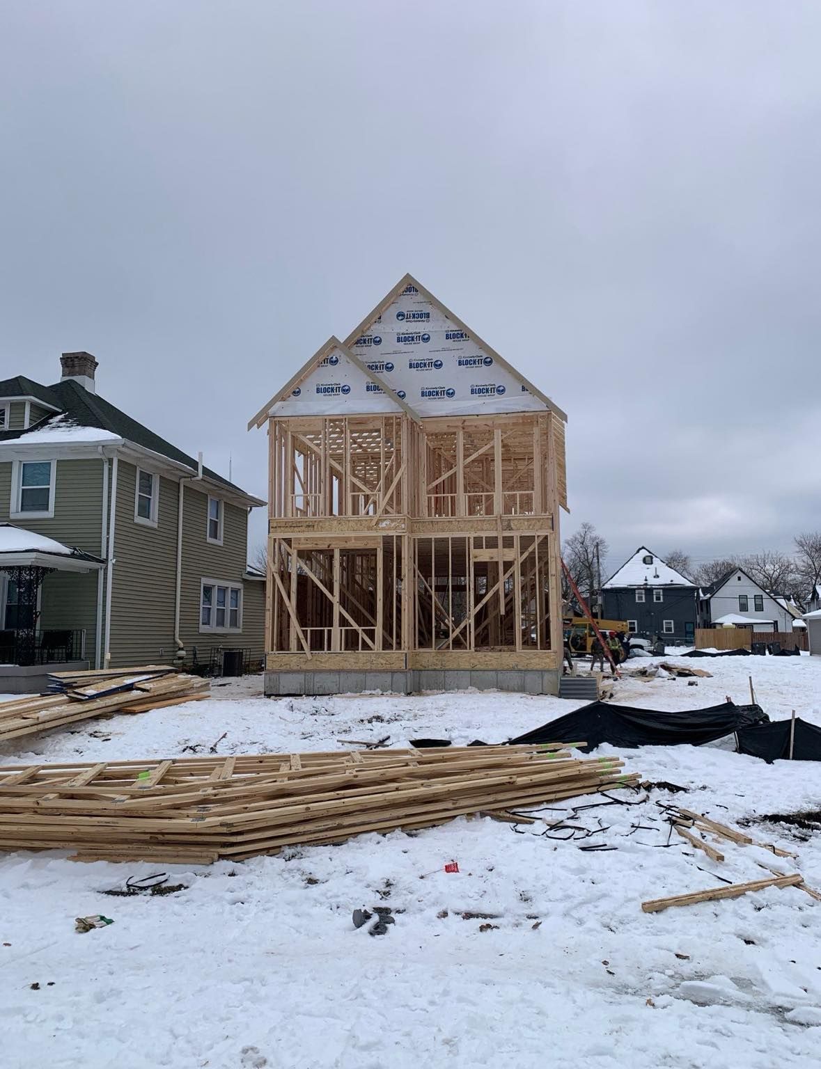 Two-story house frame under construction in snowy field, next to existing homes. Two-story house frame under construction in snowy field, next to existing homes.