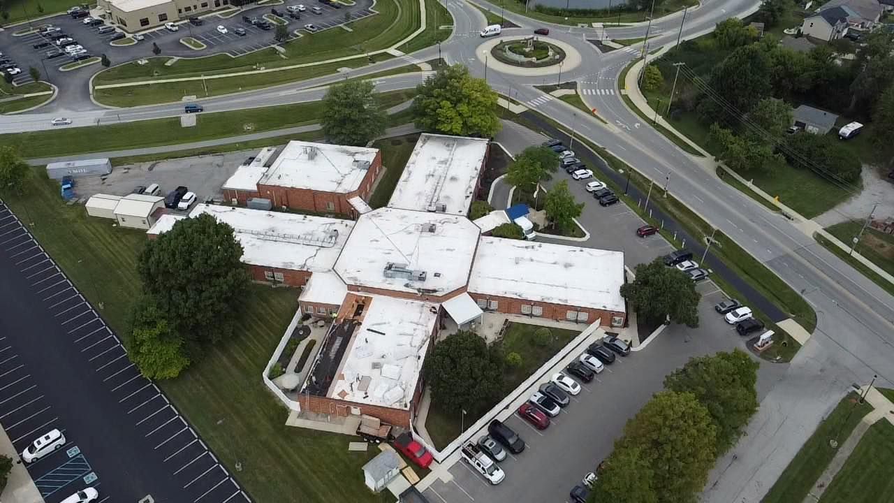 Aerial view of a multi-winged building with white roofs and a roundabout in the background. Parking lots and trees are also visible.