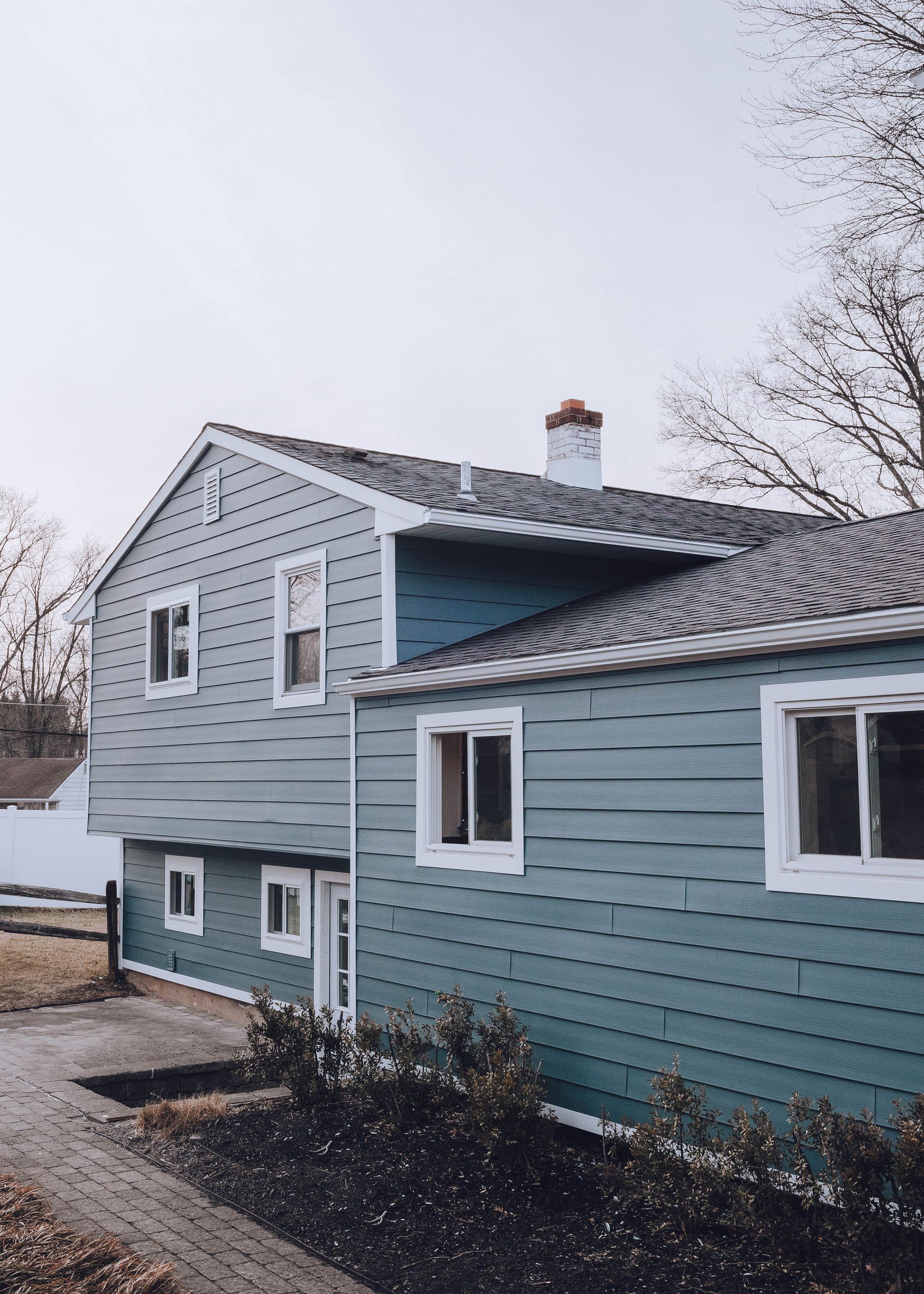 Two-story tan house with brick accents, blue shutters, and a front walkway under a cloudy sky.