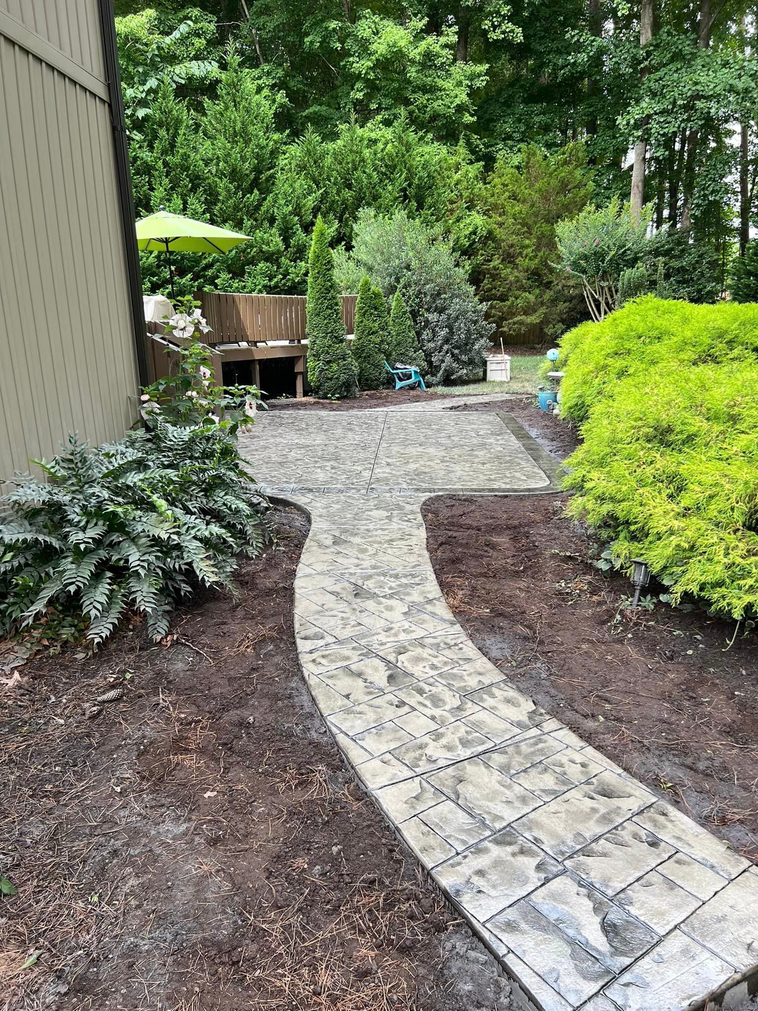 Stone pathway curves through a garden with green plants and a patio with a green umbrella.