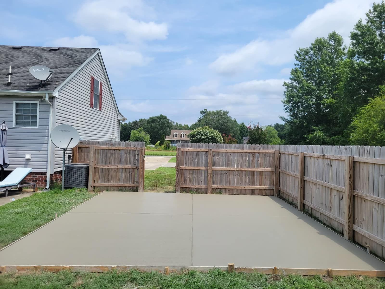 A freshly poured concrete patio enclosed by a wooden fence in a backyard, under a blue sky.