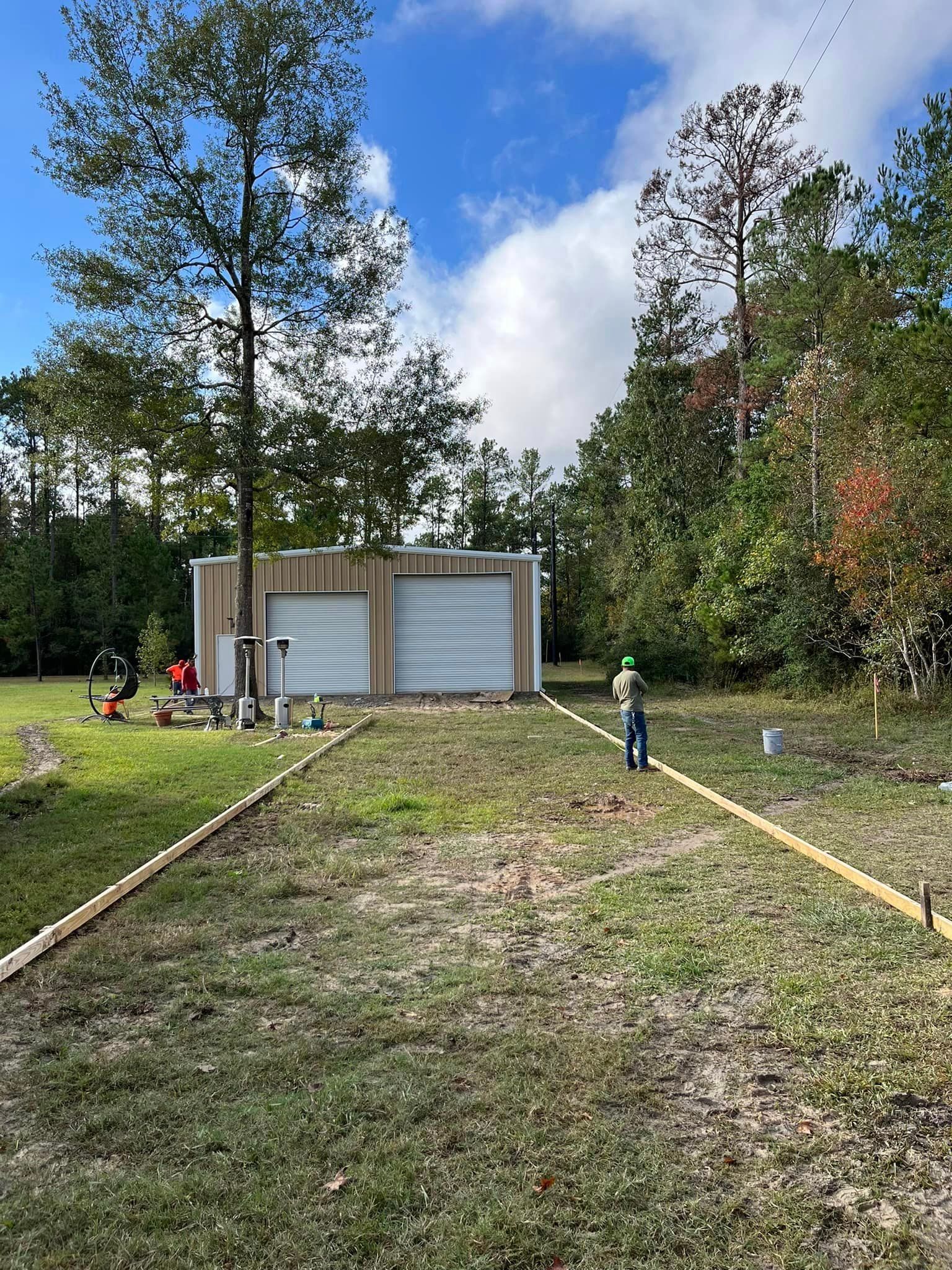 Construction site: a new garage with two bays sits in a grassy clearing; a worker stands near wood edging.