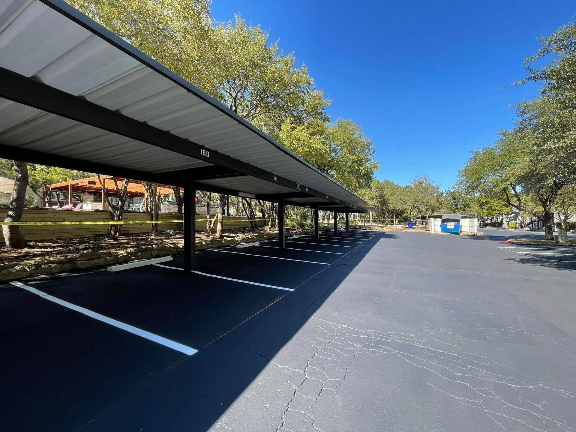 Parking lot with covered stalls, asphalt surface, trees, and blue sky.