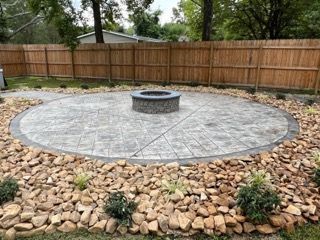 Circular stone patio with a fire pit, surrounded by rocks and a wooden fence.