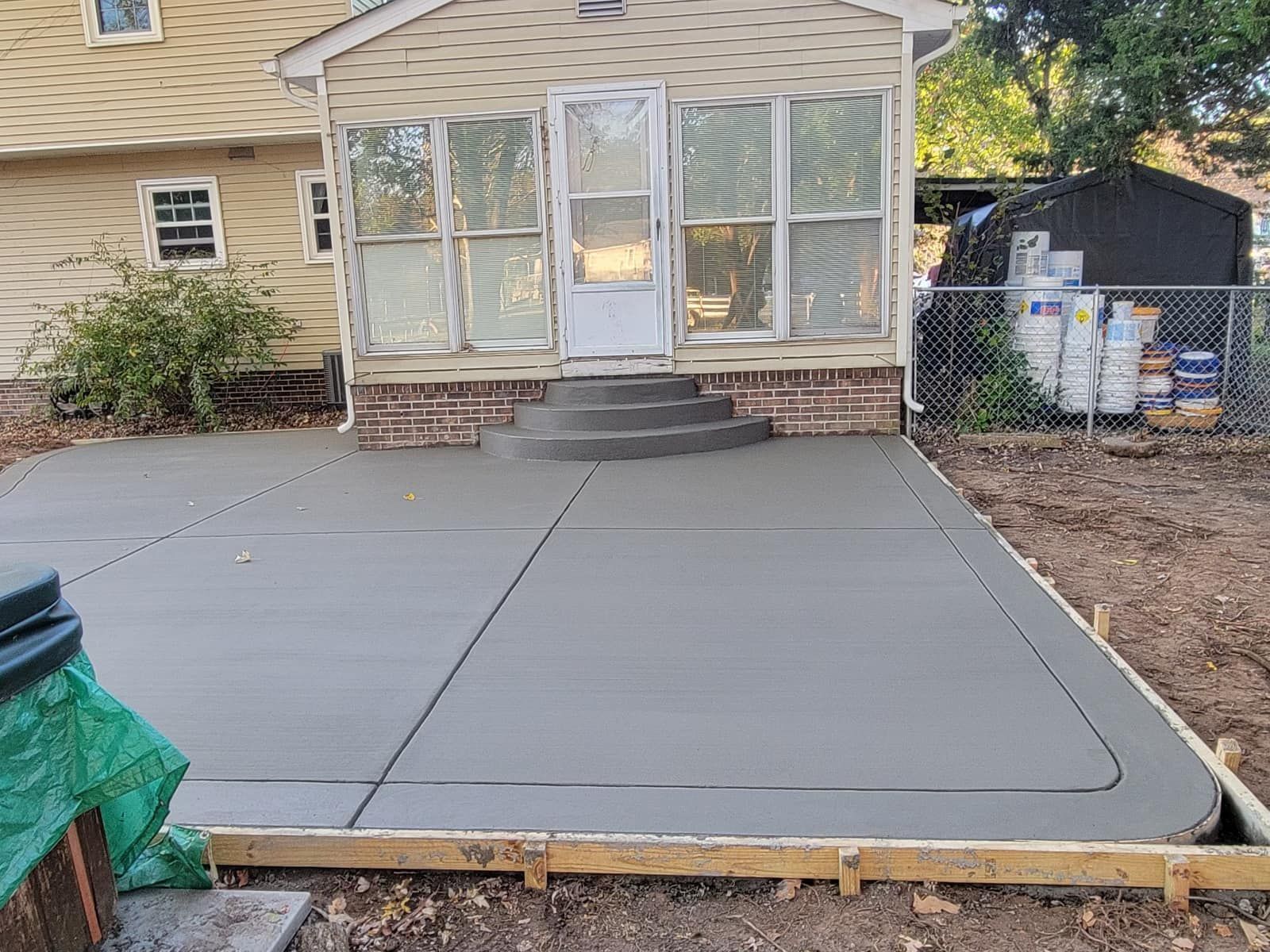 Newly poured concrete patio with a curved edge, adjacent to a house with a brick foundation.