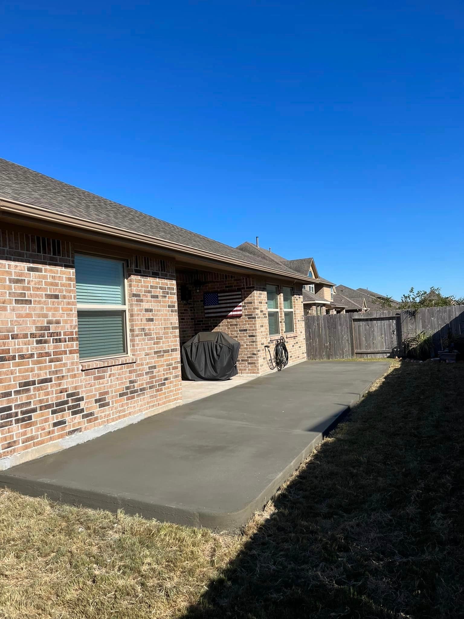 Newly poured concrete patio next to a brick house, with a grill and wooden fence on the right.