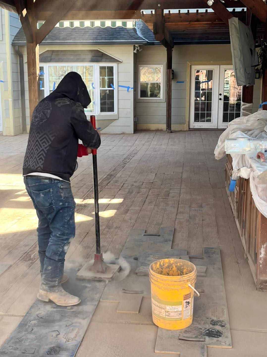 A person in a hooded jacket using a tool on paving stones outdoors, near a building with a patio.