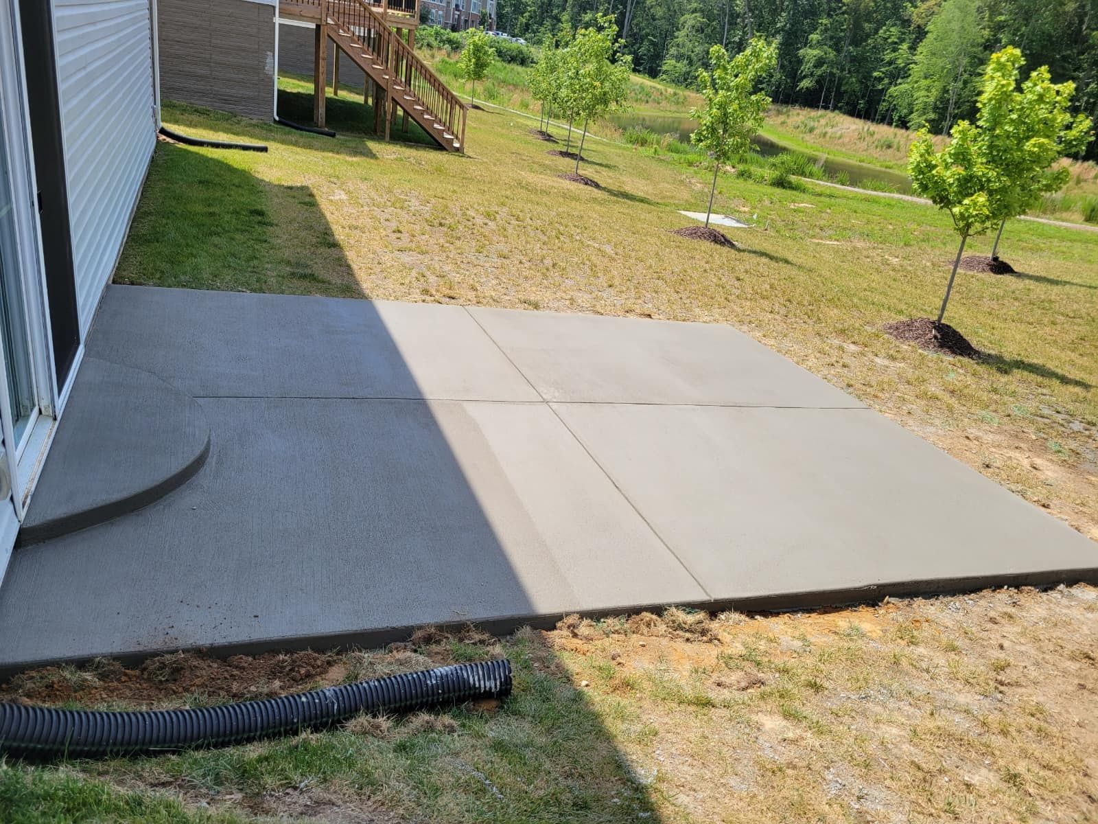 A newly poured gray concrete patio outside a house, in a grassy yard with small trees and a wooden staircase.