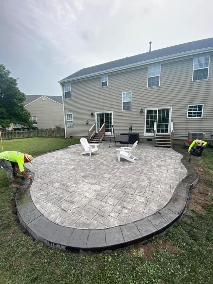 Backyard patio construction in progress; workers, grey pavers, dark border, white chairs, two-story house.