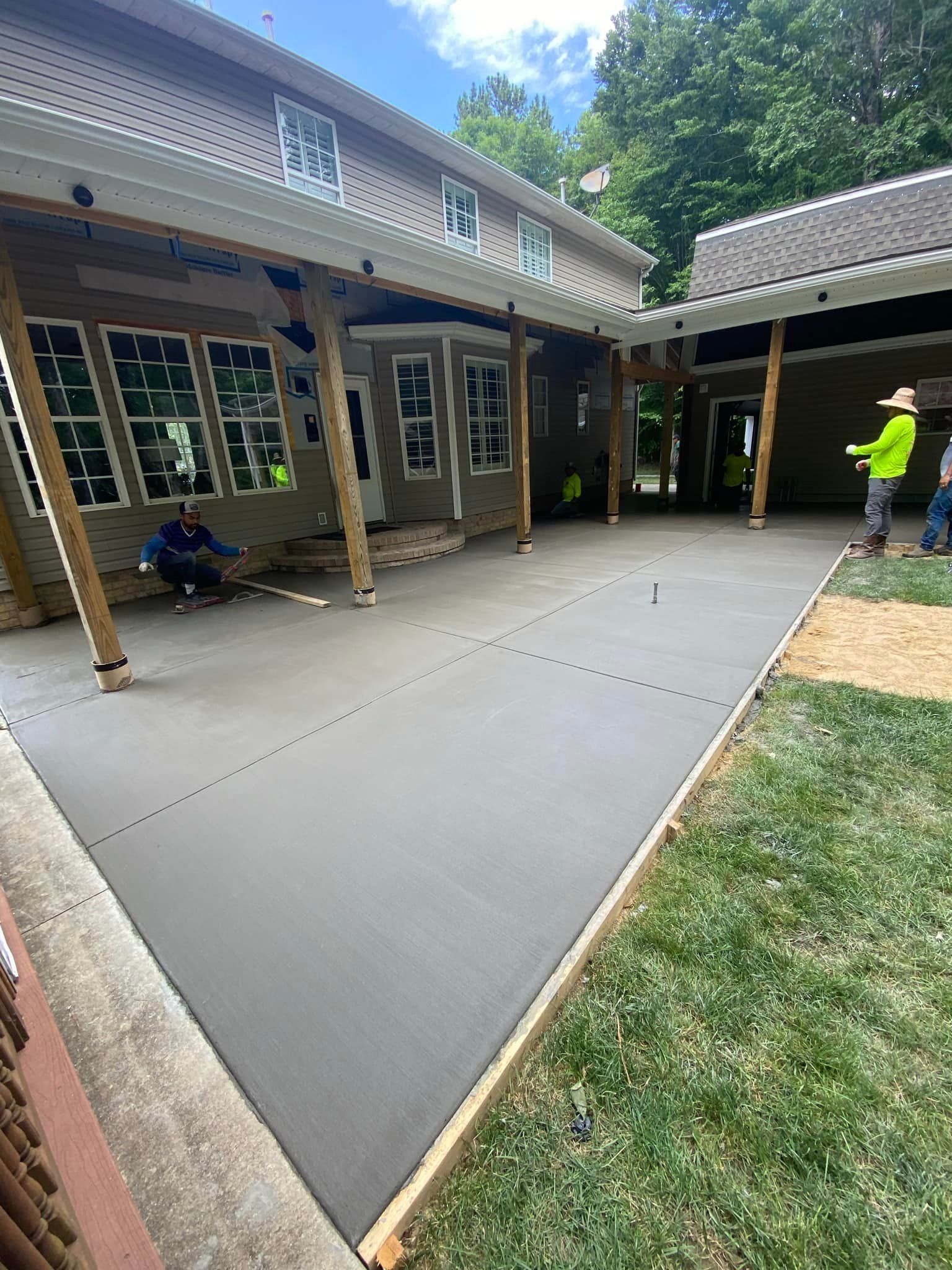 Newly poured concrete patio adjacent to a house; construction workers are finishing the surface.