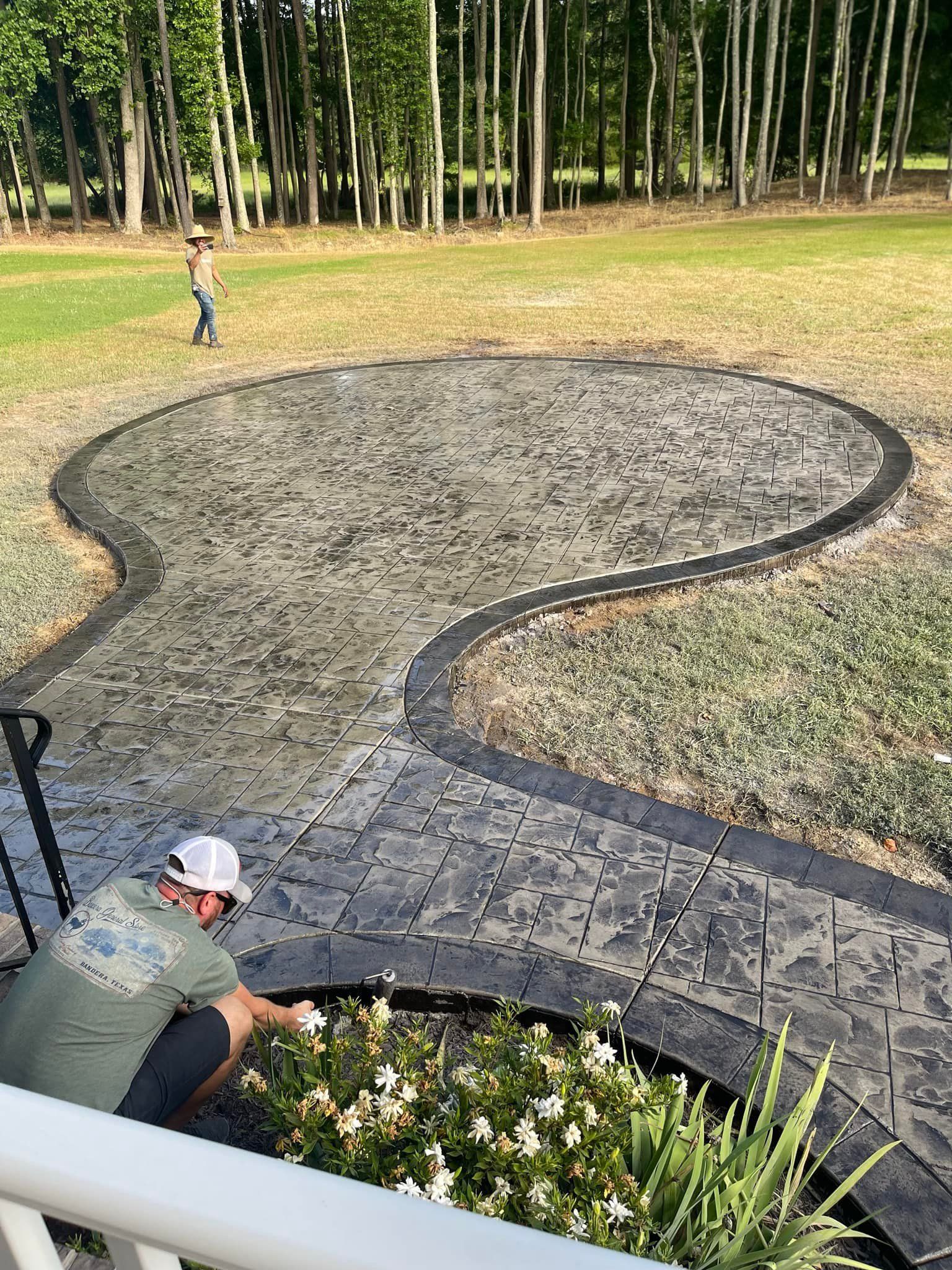 Man installing plants next to patterned concrete patio with curved walkway; person walking in background.
