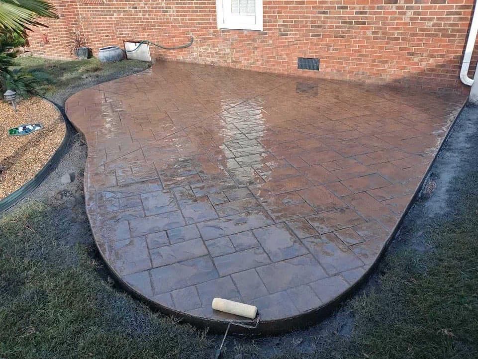 Stamped concrete patio, brick home exterior. Brown, wet surface with stone pattern, surrounded by grass.
