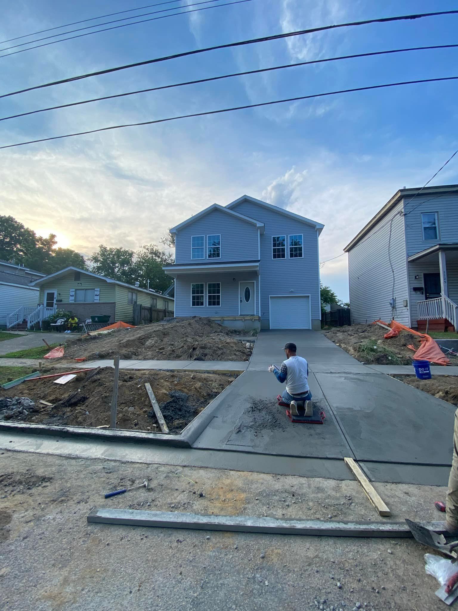 Construction worker finishing a concrete driveway in front of a two-story blue house under a partly cloudy sky.