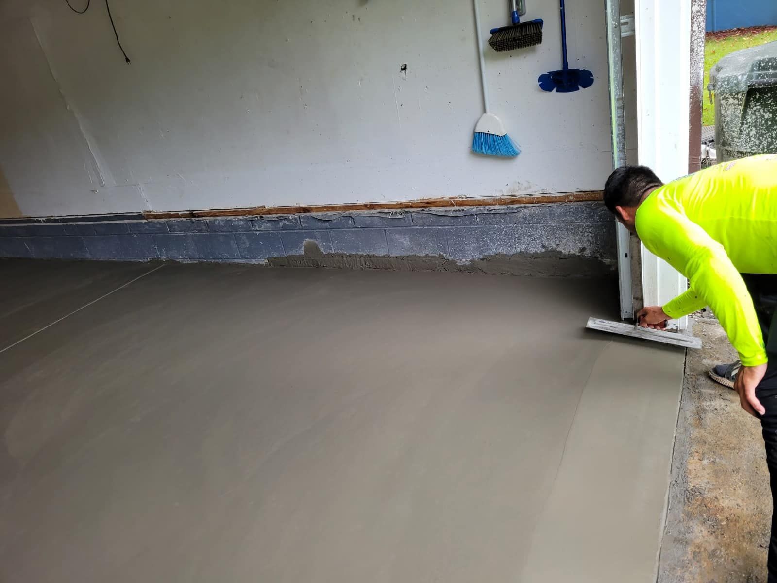 Person smoothing fresh concrete floor in a garage; wearing neon yellow shirt, using a trowel.