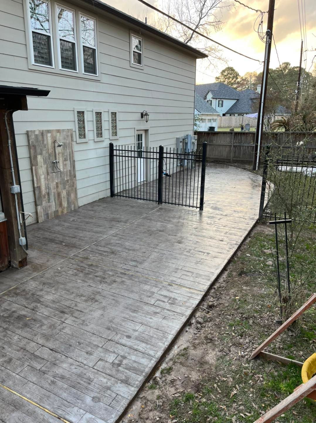 A gray stamped concrete patio, with black metal fence and wooden structure, next to a light-colored house.