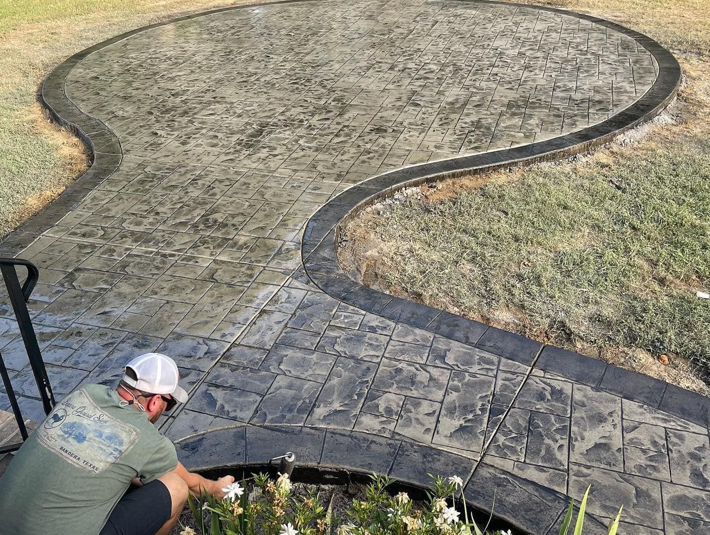 Man working on dark gray stamped concrete patio with curved border, lawn surrounds.