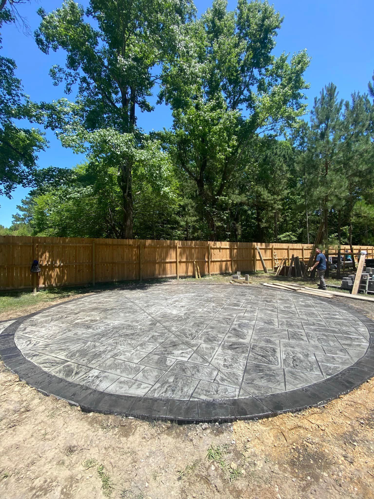 Oval paved area with black border, surrounded by dirt and wooden fence, trees in the background.