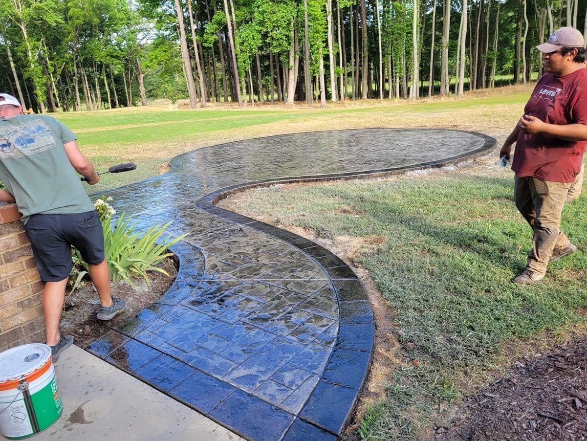 Two men applying sealant to a curved, stamped concrete walkway. Green grass and trees in the background.