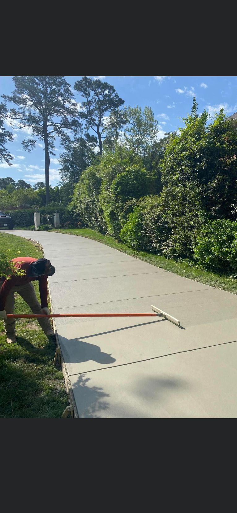 Person smoothing concrete sidewalk with a long tool; surrounded by grass and trees. Sunny day.