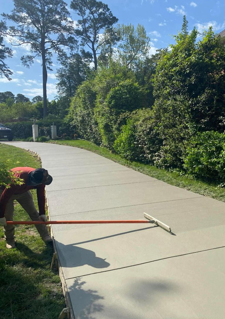 Person smoothing wet concrete driveway with a long tool. Trees and shrubs line the side.