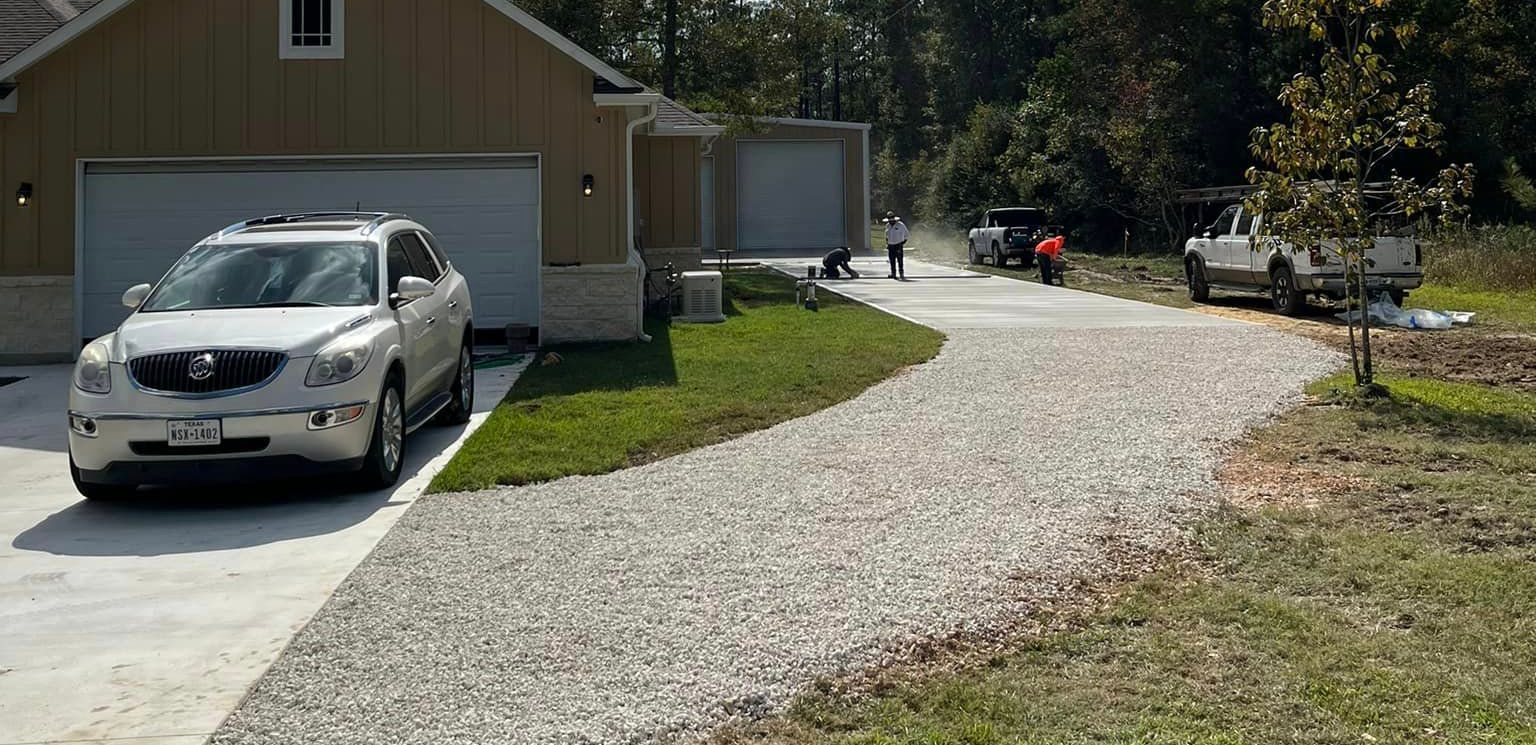 A white SUV parked in front of a garage, a gravel driveway with workers and trucks.
