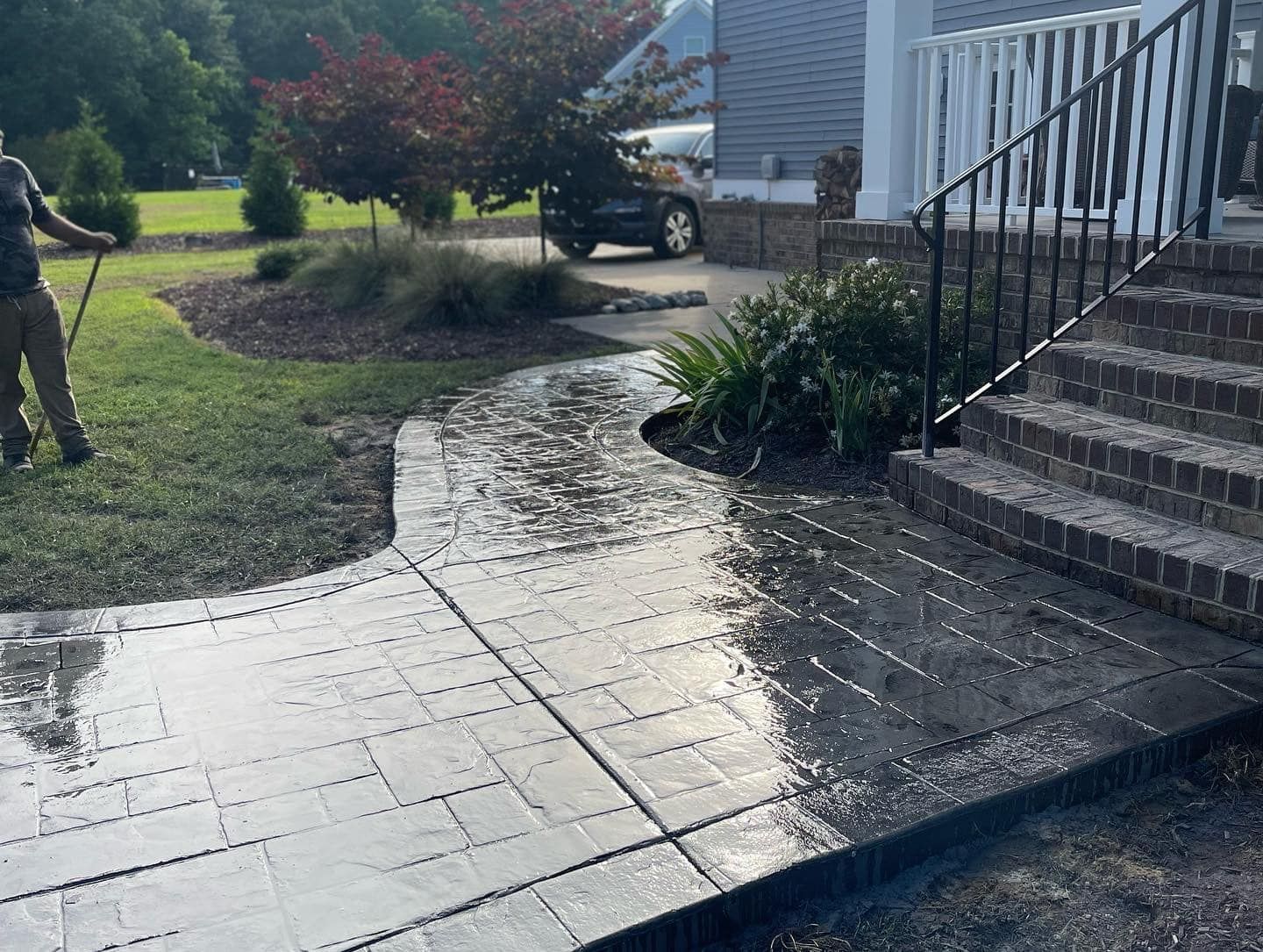 Person spraying water on wet, patterned concrete walkway leading to house steps. Green grass surrounds.